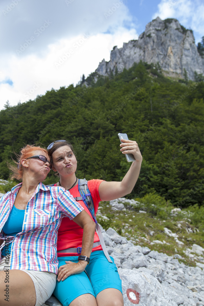 Two pretty smiling women having fun making duckface and photographing a selfie on mountain peak, whilst resting on big rock 

