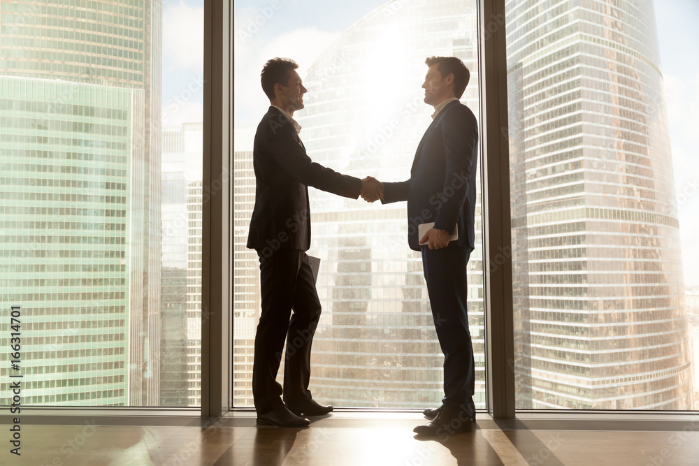 Two businessmen shaking hands standing at big window with urban cityscape, confident business partners handshaking in office, forming good relations, establish partnership, support, help. Side view Two businessmen shaking hands standing at big window with urban cityscape, confident business partners handshaking in office, forming good relations, establish partnership, support, help. Side view