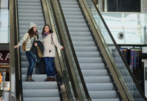 Two young women with small luggage on the escalator at the airport.