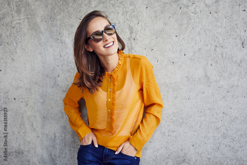 Smiling girl in sunglasses leaning against wall with hands in pockets Smiling girl in sunglasses leaning against wall with hands in pockets