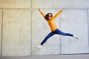 Joyful young lady jumping and raising arms in front of wall outside