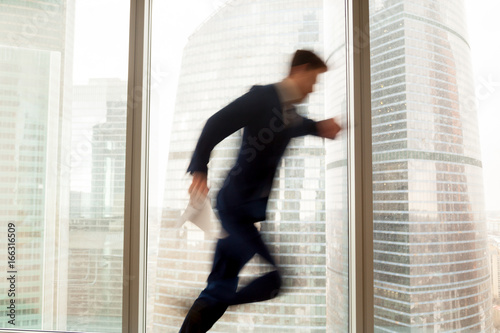 Busy businessman hurrying up to come at meeting on time in office building, blurred silhouette running in hurry along hallway, looking on wristwatch on the move, window city view at background