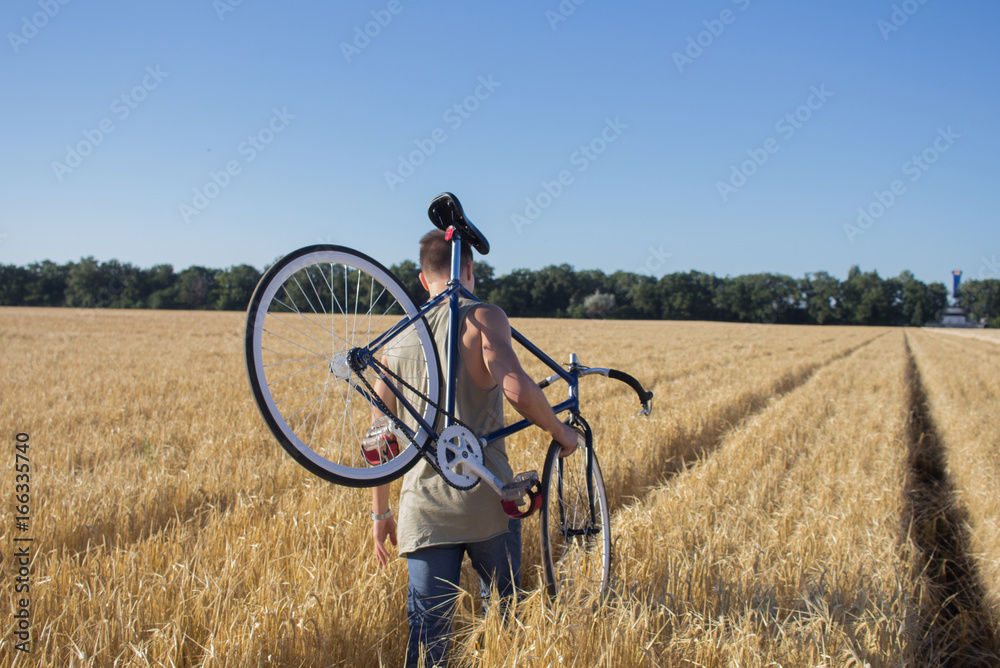 Naklejka premium young hipster in sunglasses walk in the fields with bicycle on shoulder, fields and blue sky background 