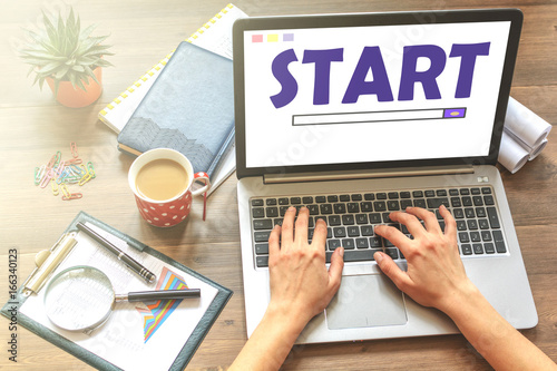 Girl is sitting at table with business accessories, notebook (diary), cup of coffee and a laptop with text Start. Top view.