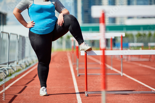 Confident and ambitious chubby female in activewear standing on racetrack and keeping one leg on hurdle