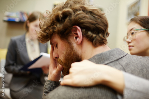 Portrait of young man crying in couples counseling session with wife hugging him supportively