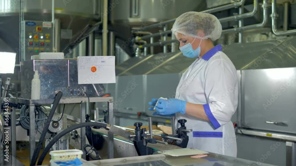 A factory operator in a plastic cap and a mask checks cheese packages. 4K. A factory operator in a plastic cap and a mask checks cheese packages. 4K.