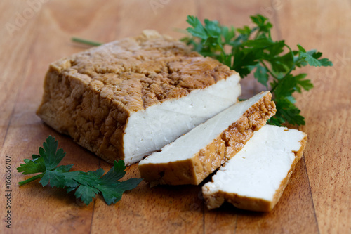 Block of smoked tofu, two tofu slices and fresh parsley on wooden chopping board.
