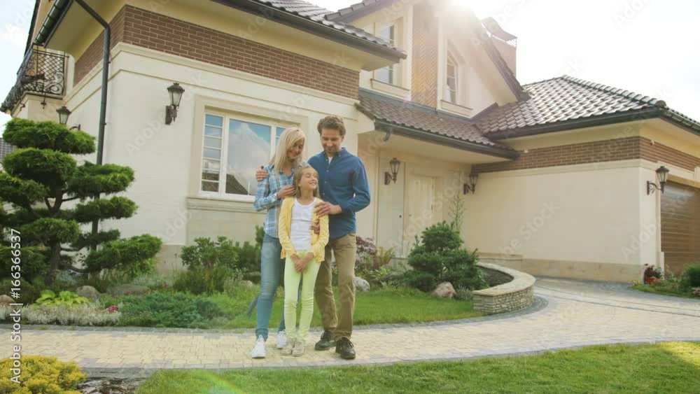 Attractive caucasian couple in front of new home during the sunset hugging their beatiful daughter and smiling on camera.