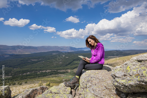 sport hiking or trekking woman with purple jacket, sitting on rock peak, watching digital tablet, next to Lozoya Valley and Guadarrama Park, in Madrid, Spain 