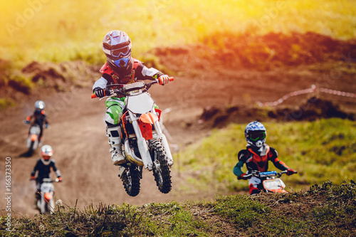 Fototapeta Young child racer on a motorcycle participates in motocross cross-country in flight, jumps and takes off on a springboard on the team of rivals