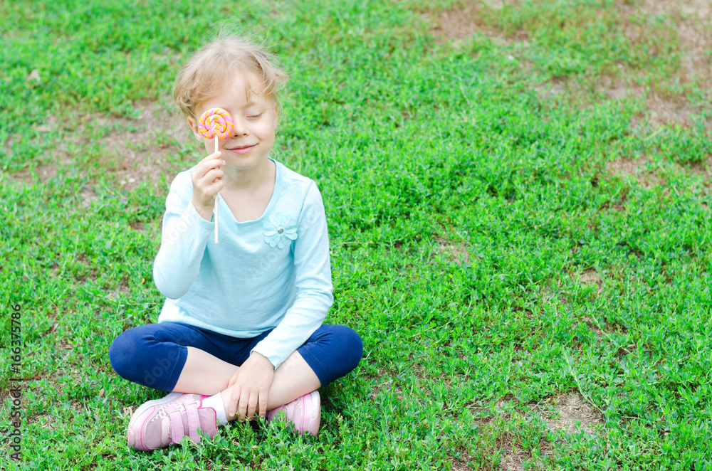 Fototapeta premium Happy little girl with a lollipop sitting on a green glade, free space for your text.
