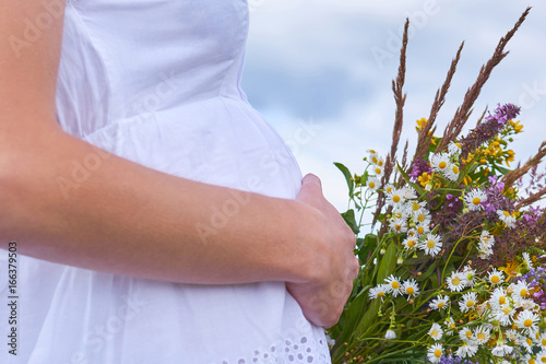Pregnant young girl stands in the field hugging her stomach by hand with a bouquet of wildflowers. The concept of motherhood and caring