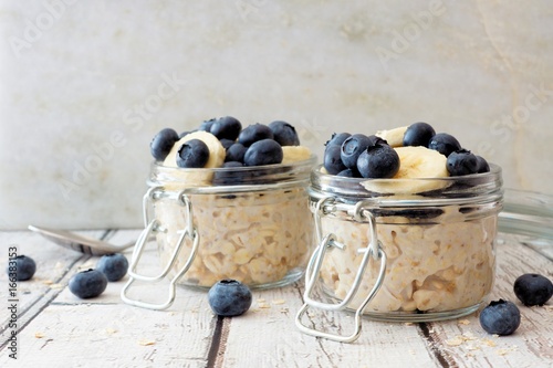 Overnight oats with fresh blueberries and bananas in jars on a rustic white wood background