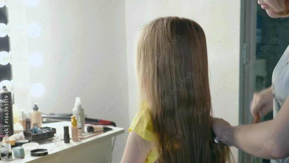 Pretty cute little girl with long beautiful hair sits in front of a mirror in a beauty salon, professional hairdresser combs her hair, use a comb. Slow motion. Side view from behind. Medium shot. Pretty cute little girl with long beautiful hair sits in front of a mirror in a beauty salon, professional hairdresser combs her hair, use a comb. Slow motion. Side view from behind. Medium shot.