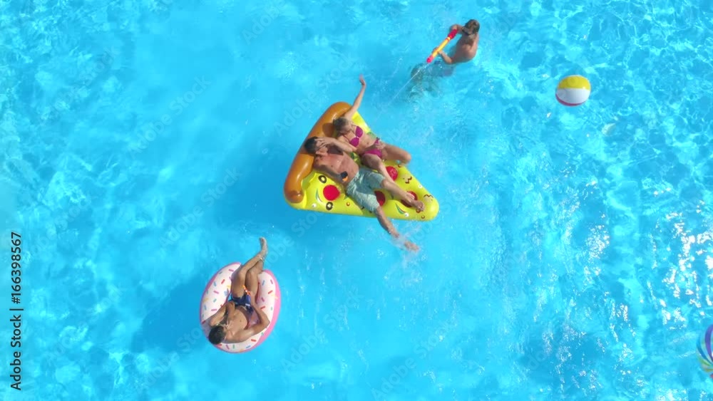 AERIAL: Cheerful friends having water fight on colorful floaties at pool party. Playful guys and girls splashing water and taking selfies on inflatable pizza, doughnut, flamingo and watermelon floats