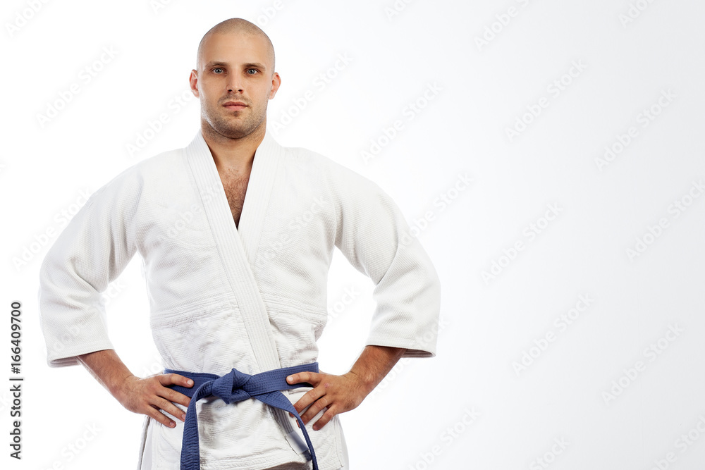 A young strong man in a white kimono for sambo, jiu jitsu and other martial  arts with a blue belt posing on white isolated background, hands holding on  his belt foto de