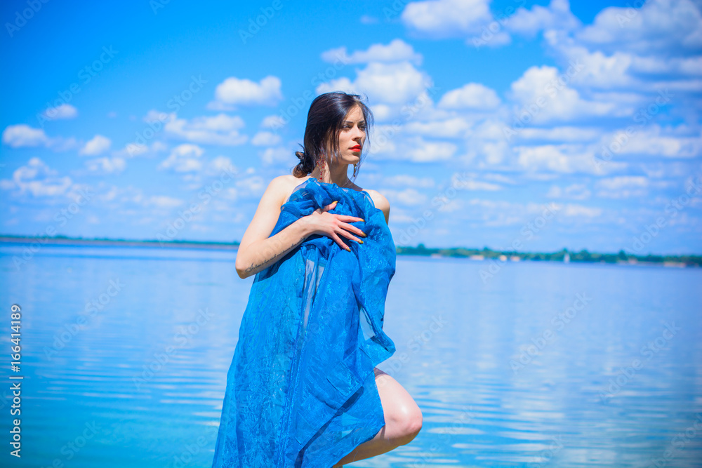 Gorgeous young brunette woman in blue fabrics, poses on the shore of the water, relaxes and shows herself in all its glory demonstrating a beautiful and well-proportioned figure Gorgeous young brunette woman in blue fabrics, poses on the shore of the water, relaxes and shows herself in all its glory demonstrating a beautiful and well-proportioned figure