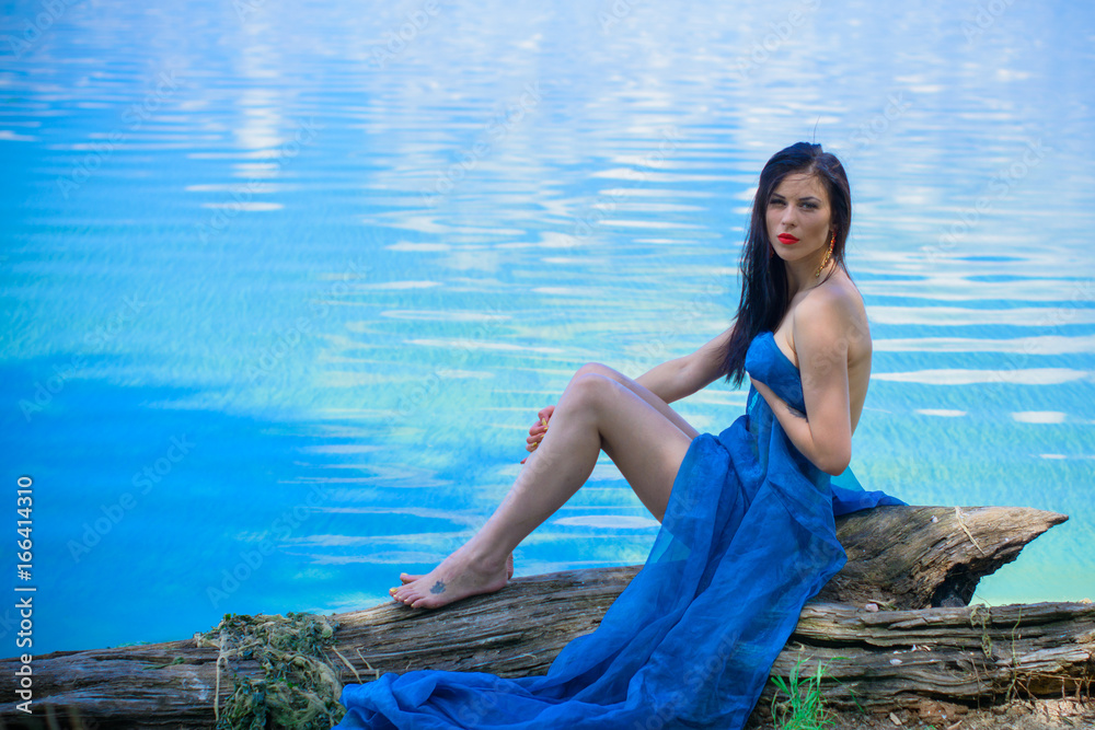 Gorgeous young brunette woman in blue fabrics, poses on the shore of the water, relaxes and shows herself in all its glory demonstrating a beautiful and well-proportioned figure