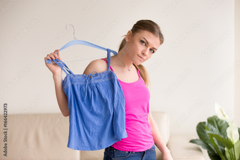 Young confused woman standing in front of mirror doubting choice of clothes for date or party, making decision what to wear, which color or style to prefer. Pensive lady holding blue blouse on hanger Young confused woman standing in front of mirror doubting choice of clothes for date or party, making decision what to wear, which color or style to prefer. Pensive lady holding blue blouse on hanger