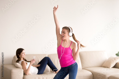 Attractive young happy lady dancing while listening her favorite song in headphones, smiling girlfriend lying in sofa nearby and using cellphone. Female friends entertain using modern digital gadgets