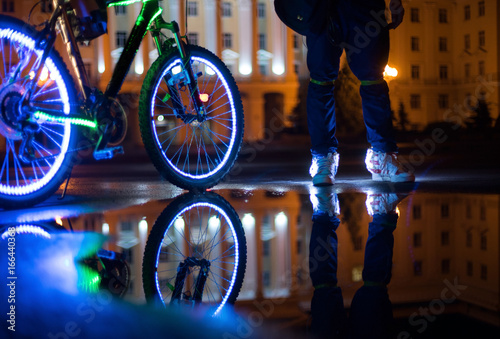 Glowing bicycle is reflected in the water. Nearby is a man in luminous shoes