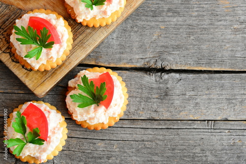 Home mini sandwiches with cream cheese on a wood background with empty place for text. Quick mini sandwiches from crackers cookies, spicy cream cheese, tomato and parsley. Tasty snack recipe. Top view