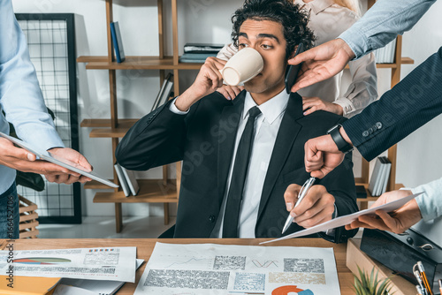 portrait of busy businessman drinking coffee and sitting at workplace while colleagues helping with work in office photo