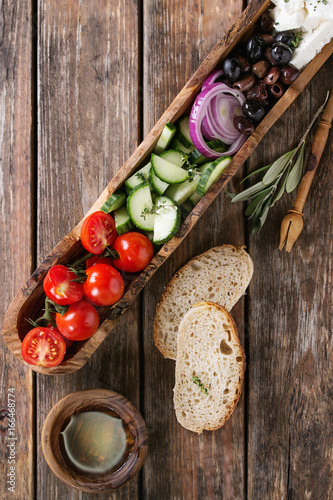 Ingredients for traditional greek salad. Cherry tomatoes, sliced cucumbers, red onion, black olives, feta cheese in olive wood bowl with loaf bread, olive oil over wooden plank background. Top view