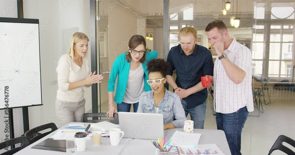 Multiracial group of young diverse people in casual clothing standing around table with coworker using laptop and watching it all together with great interest. Multiracial group of young diverse people in casual clothing standing around table with coworker using laptop and watching it all together with great interest.