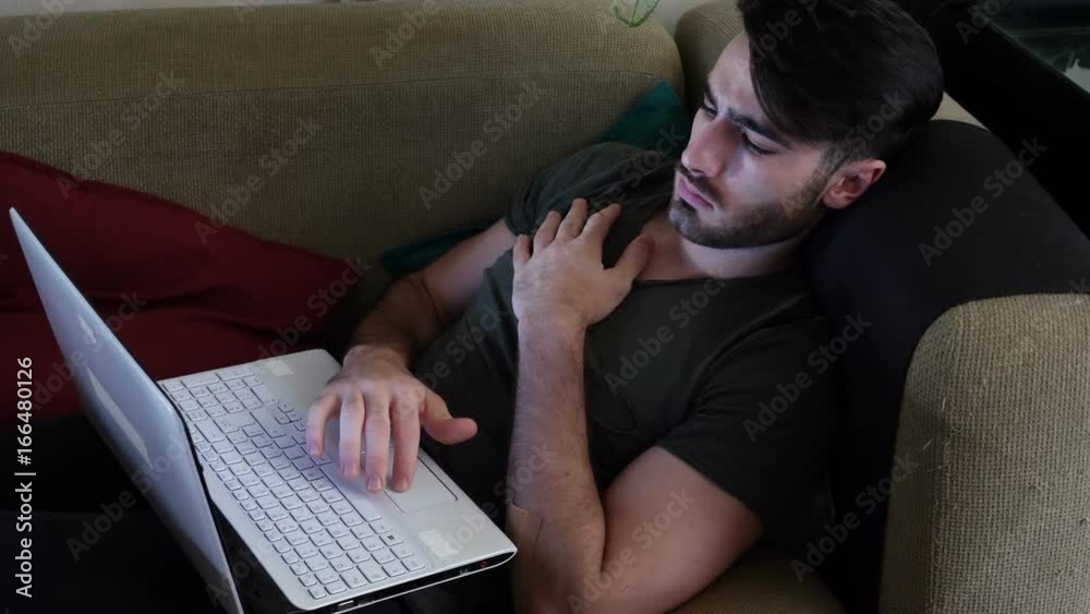Attractive Young Man with Serious Expression, with Laptop on Couch, Working on his Start-up Business - Young Male College or University Student Doing Homework on Sofa Attractive Young Man with Serious Expression, with Laptop on Couch, Working on his Start-up Business - Young Male College or University Student Doing Homework on Sofa
