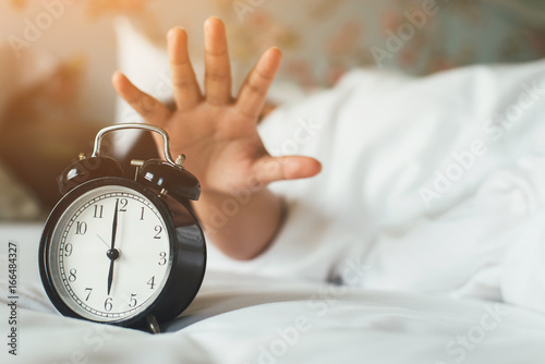 The woman's hand under the blanket reaches out for the alarm clock.