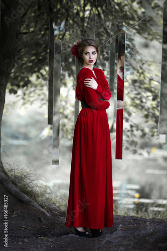 Mysterious girl in a long red dress watch mirrors in the nature.