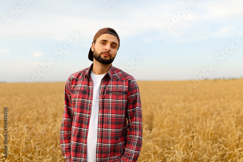 Portrait of a bearded farmer standing in a wheat field. Stilish hipster man with trucker hat and checkered shirt on. Agricultural worker