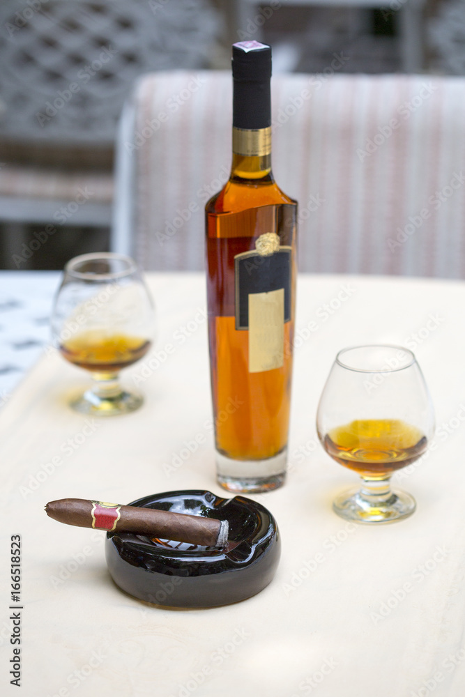 Cigar with cognac on the table in the cigar club. A box of cigars, a bottle of cognac, brandy in a glass, a sigra, a gelotine, an ashtray on a table in a restaurant.