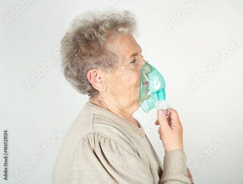 Elderly woman using asthma machine on light background
