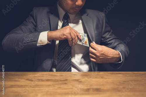 Dishonest businessman putting money, US dollars, into his suit pocket in the dark