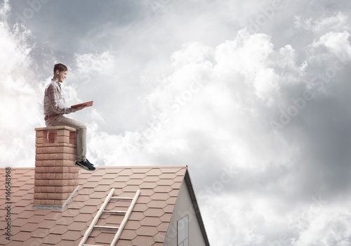 Handsome man on brick roof against cloud scape reading book