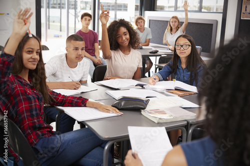 High school kids raise hands, teacher sitting at their desk © Monkey Business