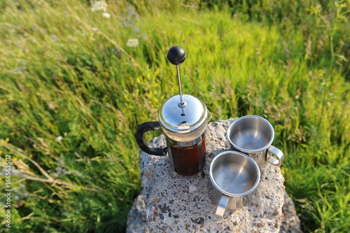 French press with coffee and two metal cups on a stone stand among summer greenery in the open air photo