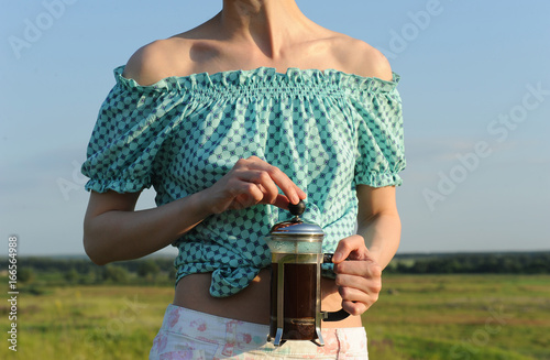 A girl is holding a French press with coffee on the background of herself in nature. Close-up photo