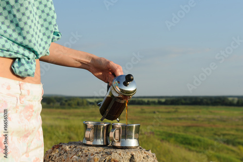 The girl pours coffee in two cups from a french-press in the open air. Summer landscape photo