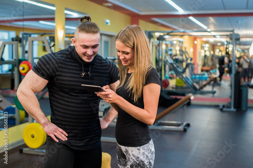 fitness, sport, exercising, technology and diet concept - smiling young woman and personal trainer with smartphone in gym
