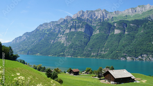 Bauernhaus am See/Blick auf den Bergsee Walensee in der Schweiz; die steilen Felswände der Churfirsten und türkisfarbiges Wasser; im Vordergrund eine Wiese mit einem Bauernhaus