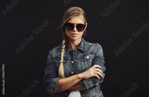 Fashion model in sunglasses with pigtail wearing trendy clothes posing in studio over black background. 