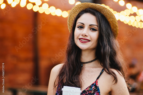 Pleased female with black hair, shining eyes and red lips, wearing straw hat and dress, holding takeaway coffee while standing at outdoor cafe resting after her working day, posing at camera