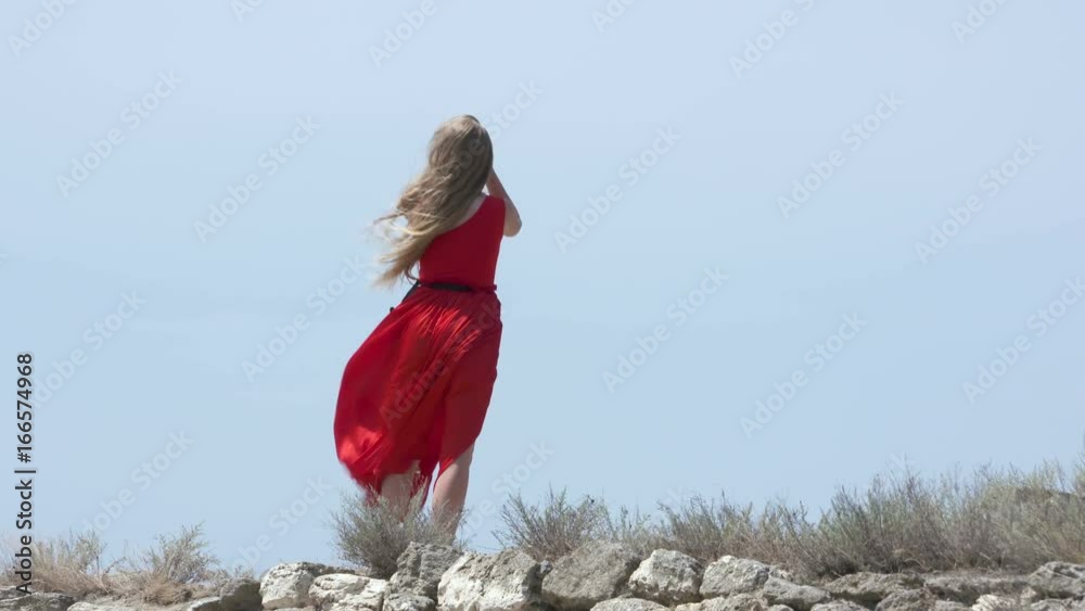 Young barefoot girl in long red dress on an old stone wall against blue sky. Red dress blowing in the wind in slow motion.