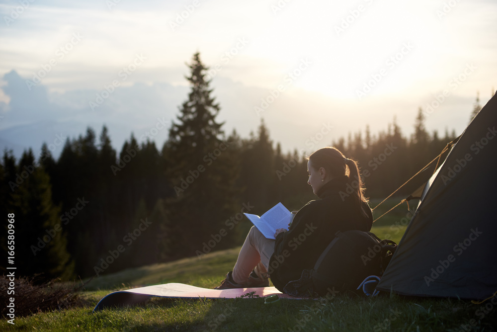 Young woman hiker reading a book near the tent in the evening at sunset