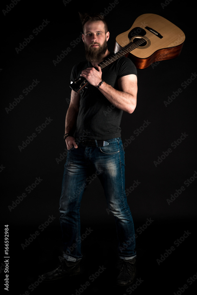 A charismatic and stylish man with a beard stands full-length with an acoustic guitar on a black isolated background.