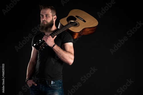 A charismatic and stylish man with a beard stands full-length with an acoustic guitar on a black isolated background.